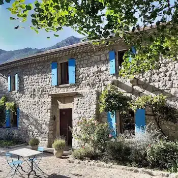 Grande maison traditionnelle en pierre en Ardèche avec volets bleus, terrasse pavée et jardin fleuri au pied des montagnes pour un séjour insolite nature.