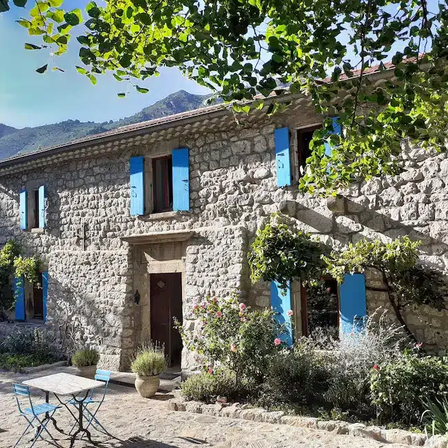 Grande maison traditionnelle en pierre en Ardèche avec volets bleus, terrasse pavée et jardin fleuri au pied des montagnes pour un séjour insolite nature.